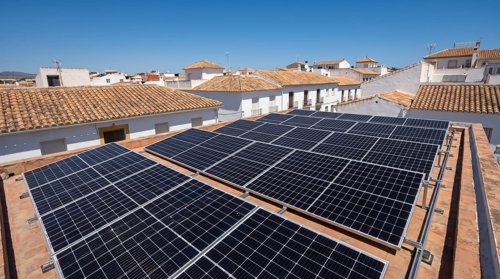 Placas solares fotovoltaicas en el tejado de una casa de un pueblo mediterráneo del sur de España, bajo el sol alto del mediodía y cielo azul, con tejados de teja al fondo.
