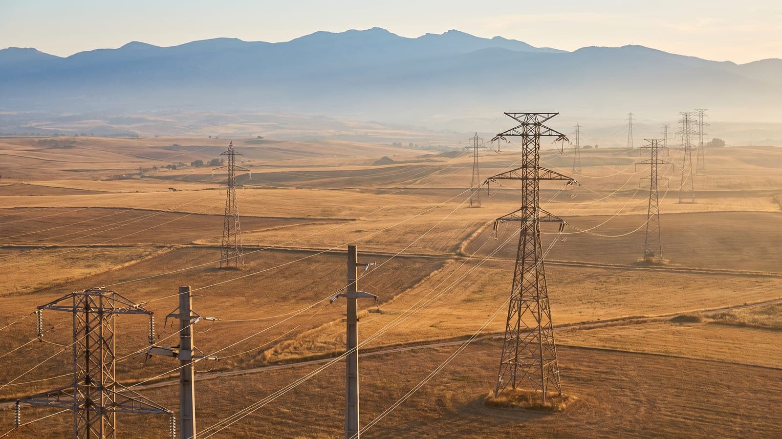 Torres de alta tensión y líneas eléctricas cruzando la meseta castellana dorada al atardecer, con montañas al fondo.