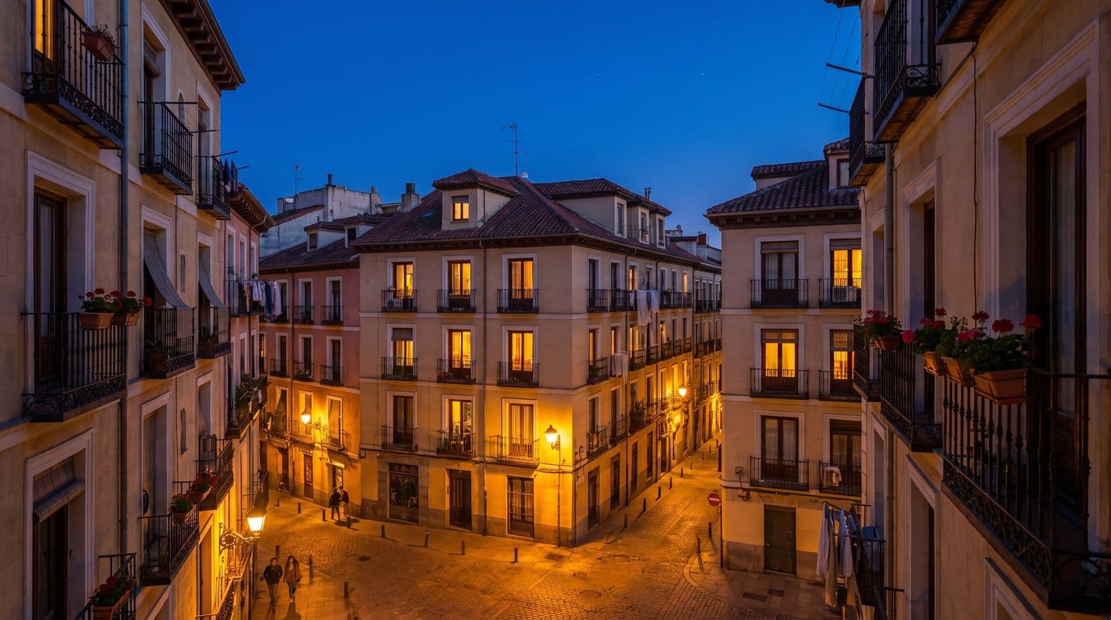 Fachadas de un barrio residencial español al anochecer con decenas de ventanas iluminadas en amarillo cálido, la hora de la cena, cuando el consumo eléctrico de los hogares llega a su punto más alto.