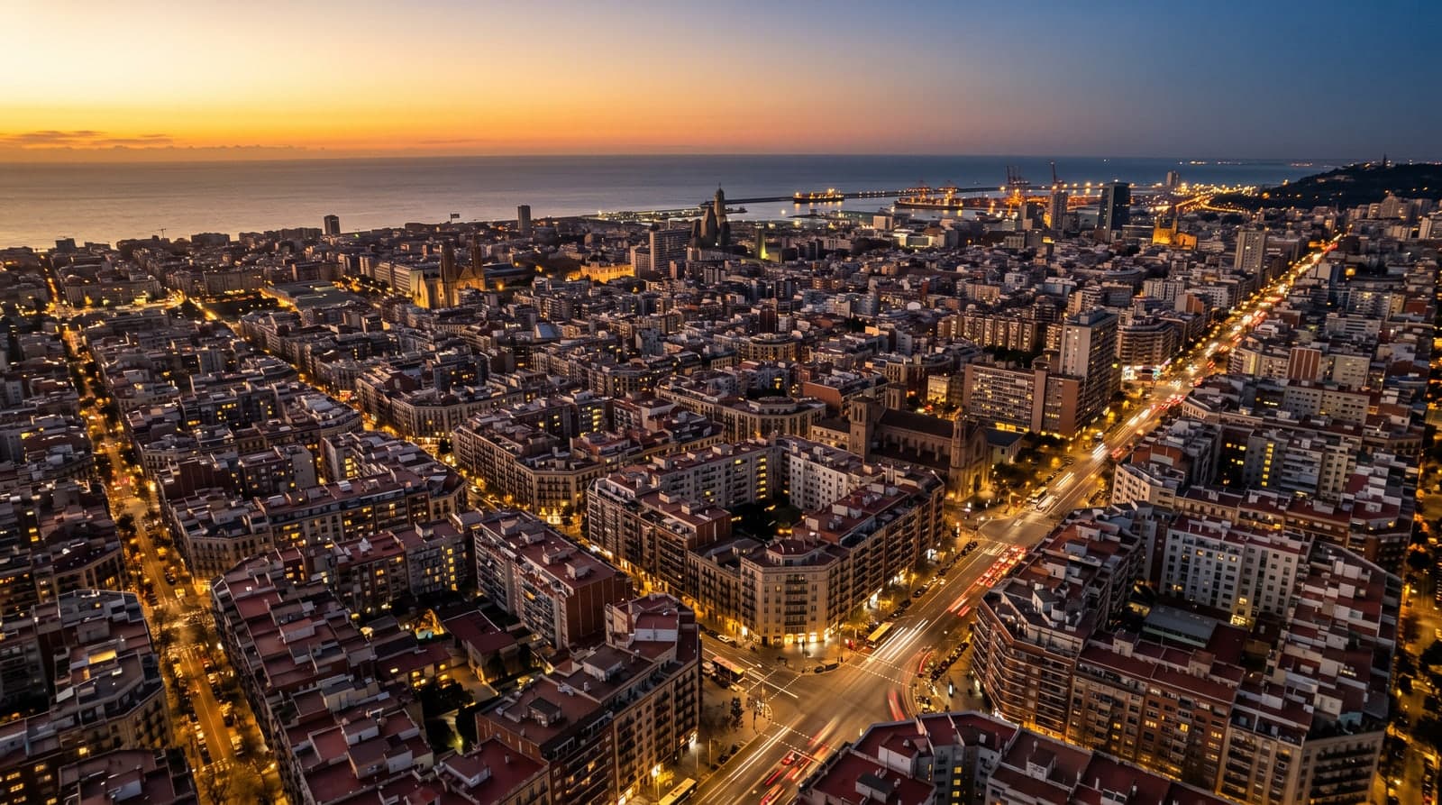 Vista aérea de una ciudad mediterránea española al anochecer, con el cielo del atardecer virando de dorado a azul mientras se encienden las luces de miles de ventanas, farolas y las estelas del tráfico en las avenidas, y el puerto y el mar al fondo.