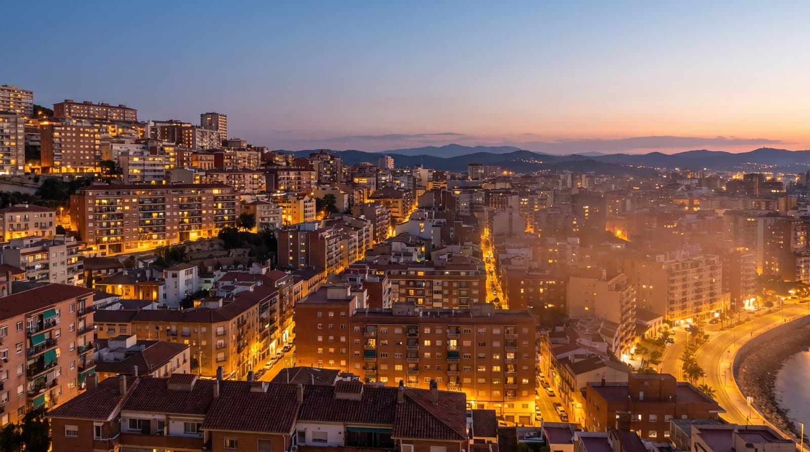 Vista aérea de una ciudad costera española al anochecer, en la hora azul, con las luces de los edificios y las farolas encendiéndose sobre las colinas y el mar al fondo.
