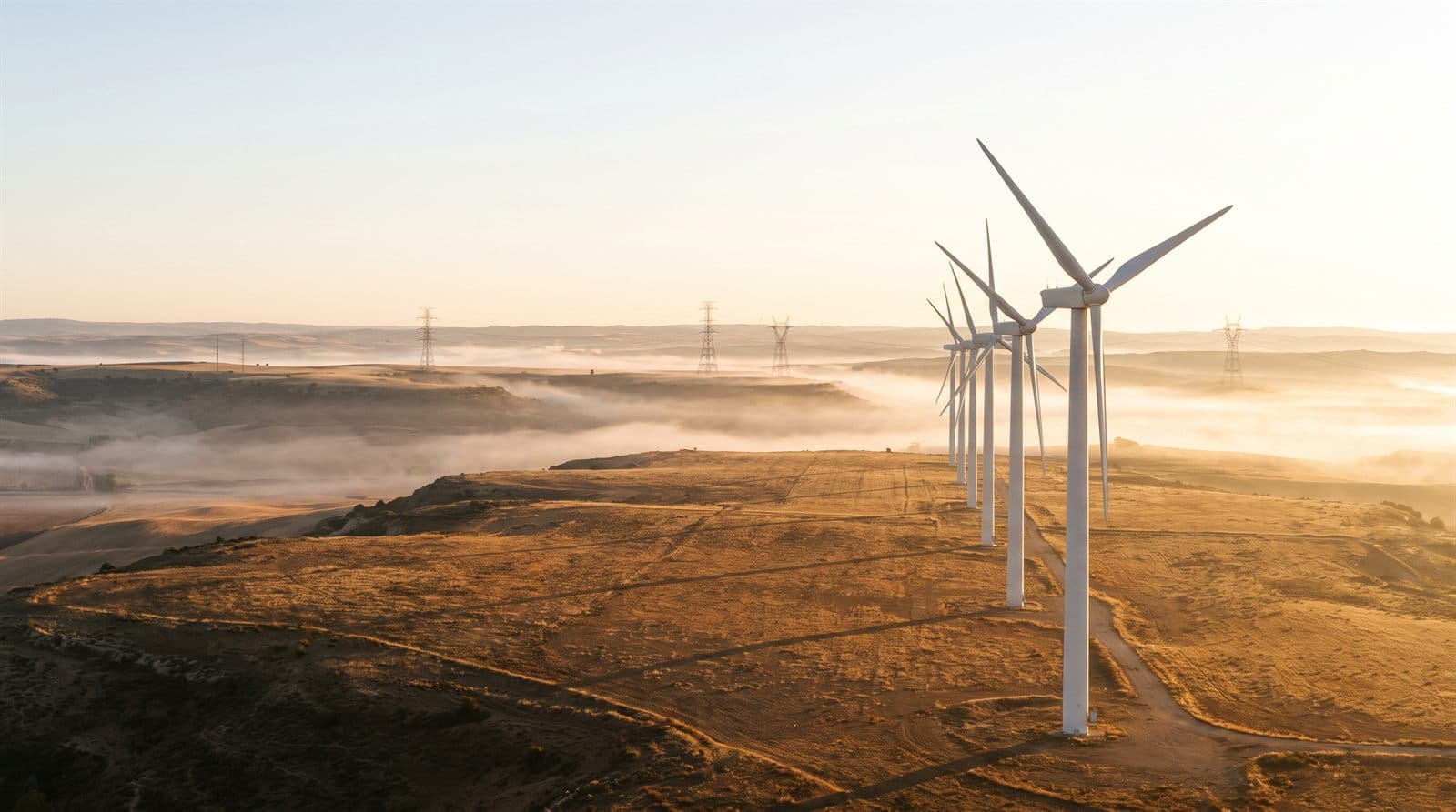 Aerogeneradores blancos alineados en una meseta de Castilla al amanecer, con niebla en el valle y torres de alta tensión al fondo