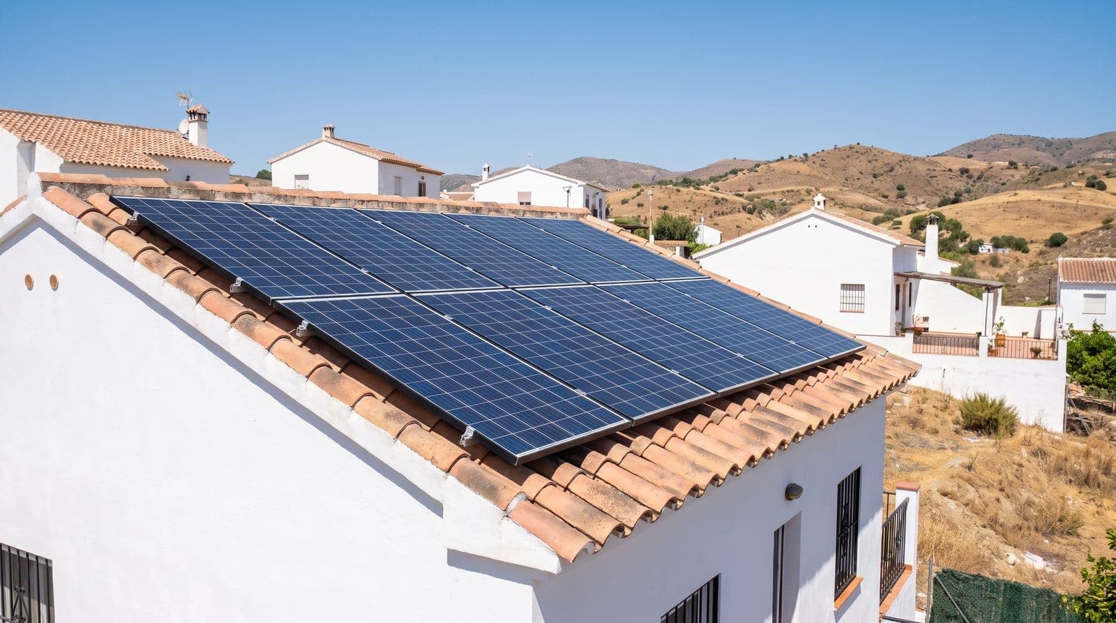 Paneles solares fotovoltaicos sobre el tejado de teja de una casa blanca mediterránea del sur de España a pleno sol del mediodía, con el cielo despejado y colinas secas al fondo.