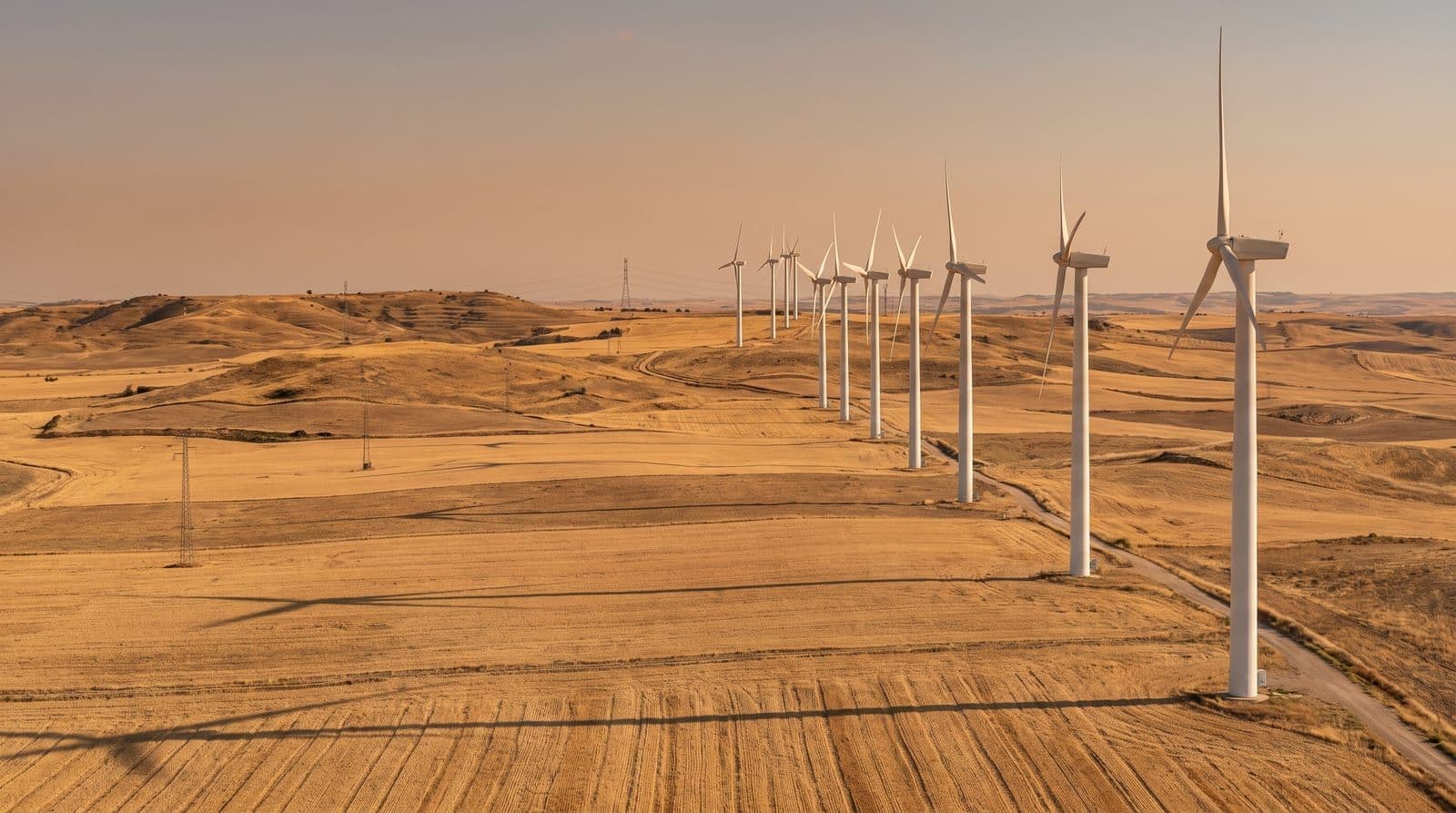 Hilera de aerogeneradores blancos sobre una meseta castellana de campos de cereal segados y colinas secas, bajo la luz cálida y dorada de la tarde de finales de verano en el interior de España.