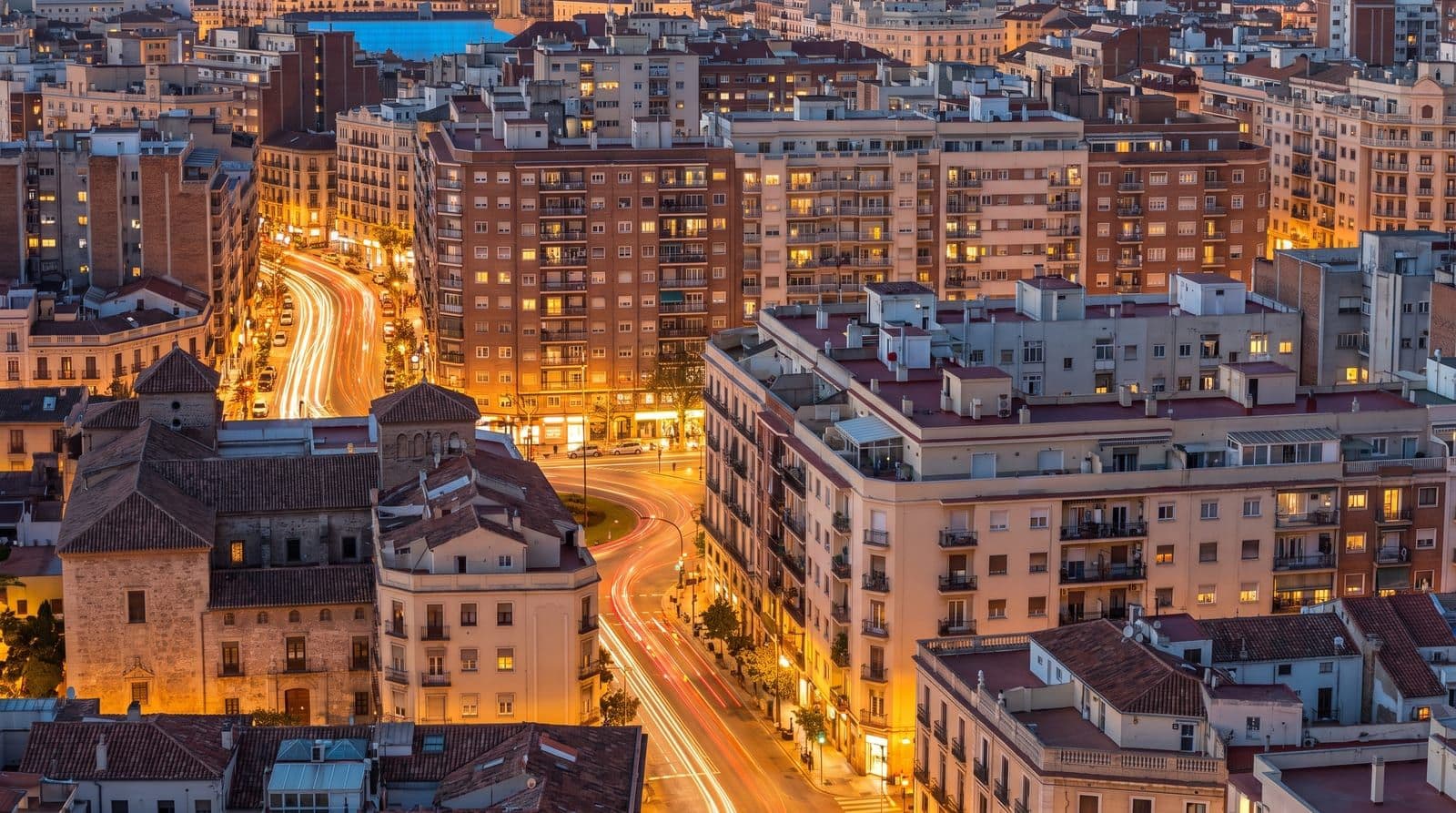 Perfil de una ciudad española al anochecer en hora punta, con las ventanas de los edificios iluminadas y las calles encendidas bajo el último resplandor azul del cielo.