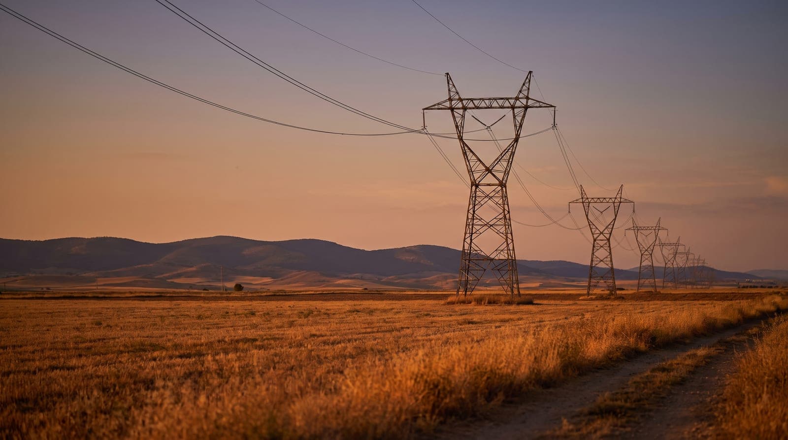 Torres de alta tensión y líneas eléctricas cruzando el campo español al atardecer, con el sol bajo en el horizonte.