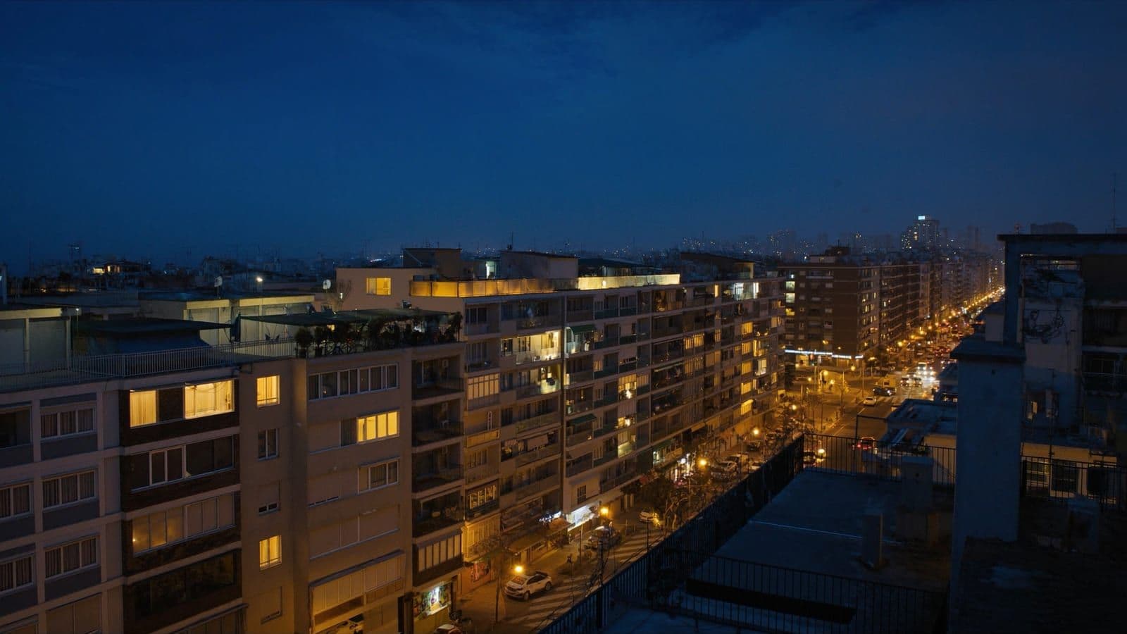 Ciudad española al anochecer, con las ventanas de los bloques de viviendas encendidas y el tráfico en la avenida durante la hora punta de la noche.