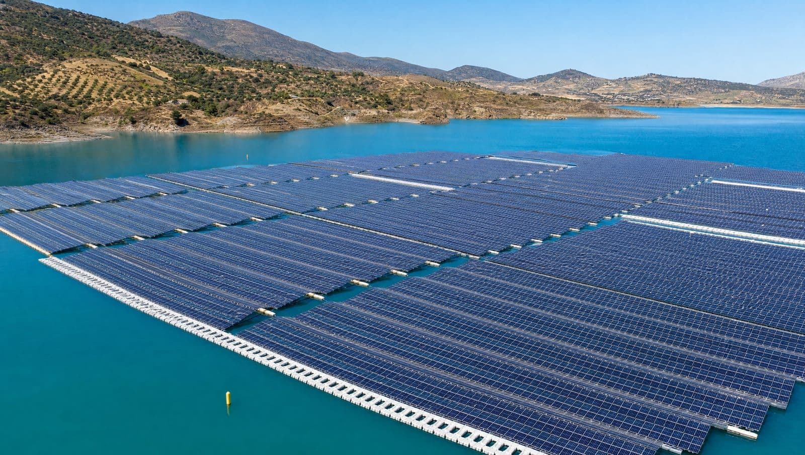 Planta solar fotovoltaica flotante sobre un embalse español al mediodía: largas filas de paneles azul oscuro apoyados en pontones sobre agua turquesa en calma, con colinas mediterráneas secas al fondo bajo un cielo despejado.