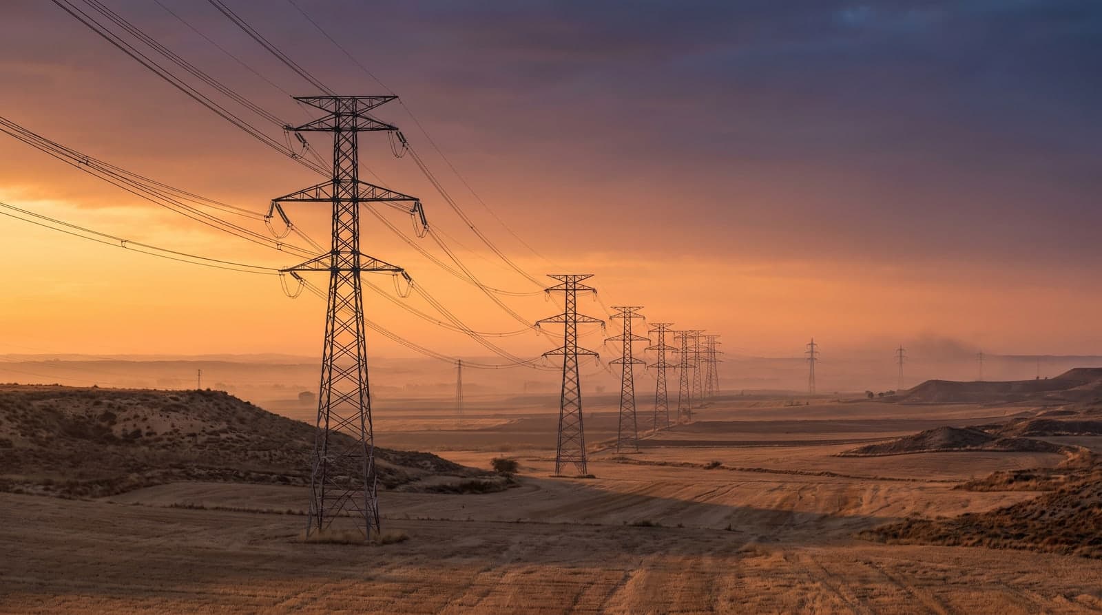Hilera de torres metálicas de alta tensión cruzando una llanura castellana seca al atardecer, con los cables recortados sobre un cielo naranja y violeta y campos de rastrojo al fondo.