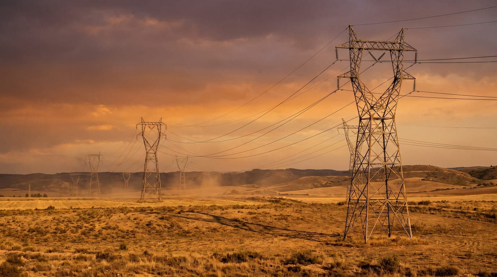 Torres de alta tensión y líneas eléctricas cruzando una meseta española seca al atardecer, bajo un cielo anaranjado de ocaso.